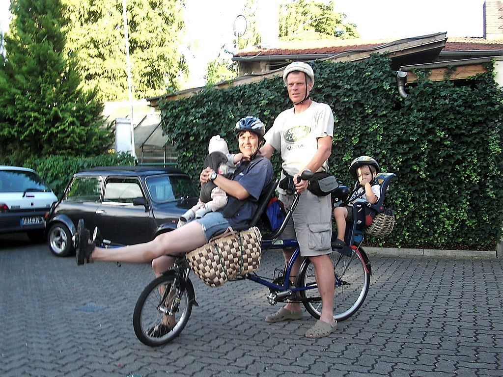 Un homme et une femme avec leur vélo en face d'une maison. Il y a un enfant qui assoit dans un siège d'enfant au-dessus le pneu derrière. La femme tient un autre enfant et les deux sont assis sur l'autre pneu. L'homme s'apprête de faire du vélo.