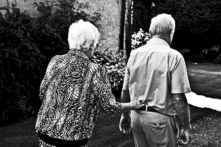 Une femme et un homme très âgés se promenade en plein air. Les deux s'éloignent de l'appareil et la femme tient la ceinture de l'homme au bas du dos.