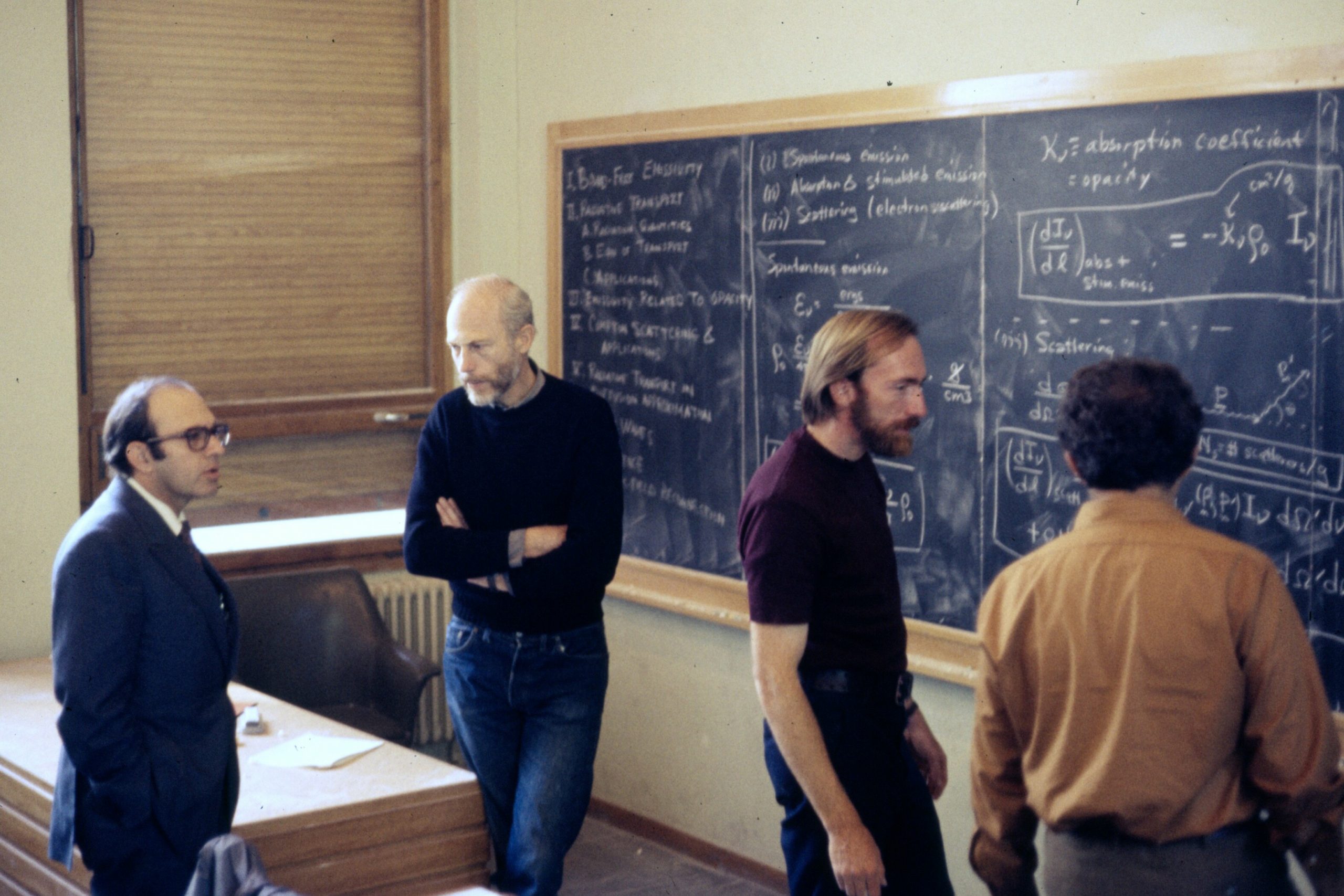 Quatre hommes dans une salle de classe universitaire qui se parlent. Ils se tiennent début en face d'un tableau.