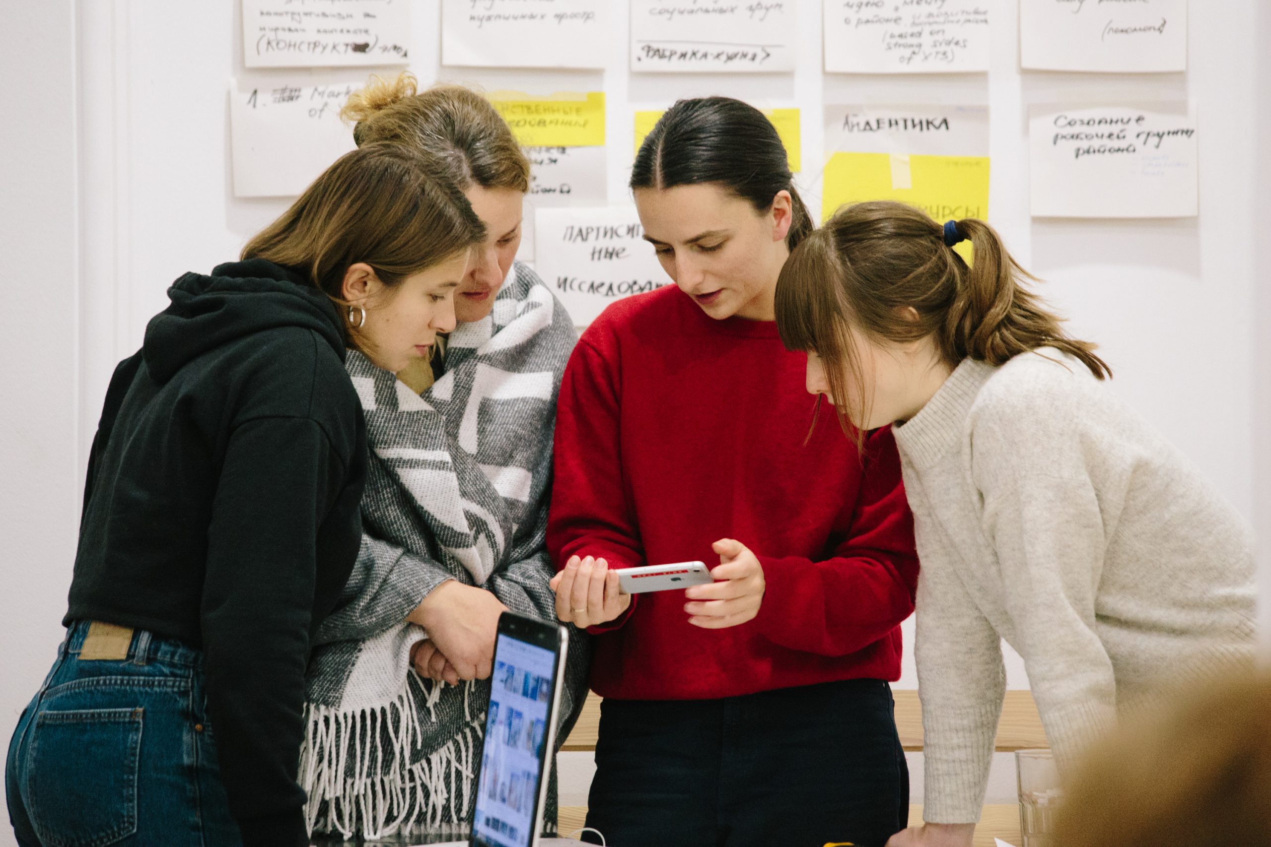 Four female students in a classroom looking at a cellphone in a semi-circle.