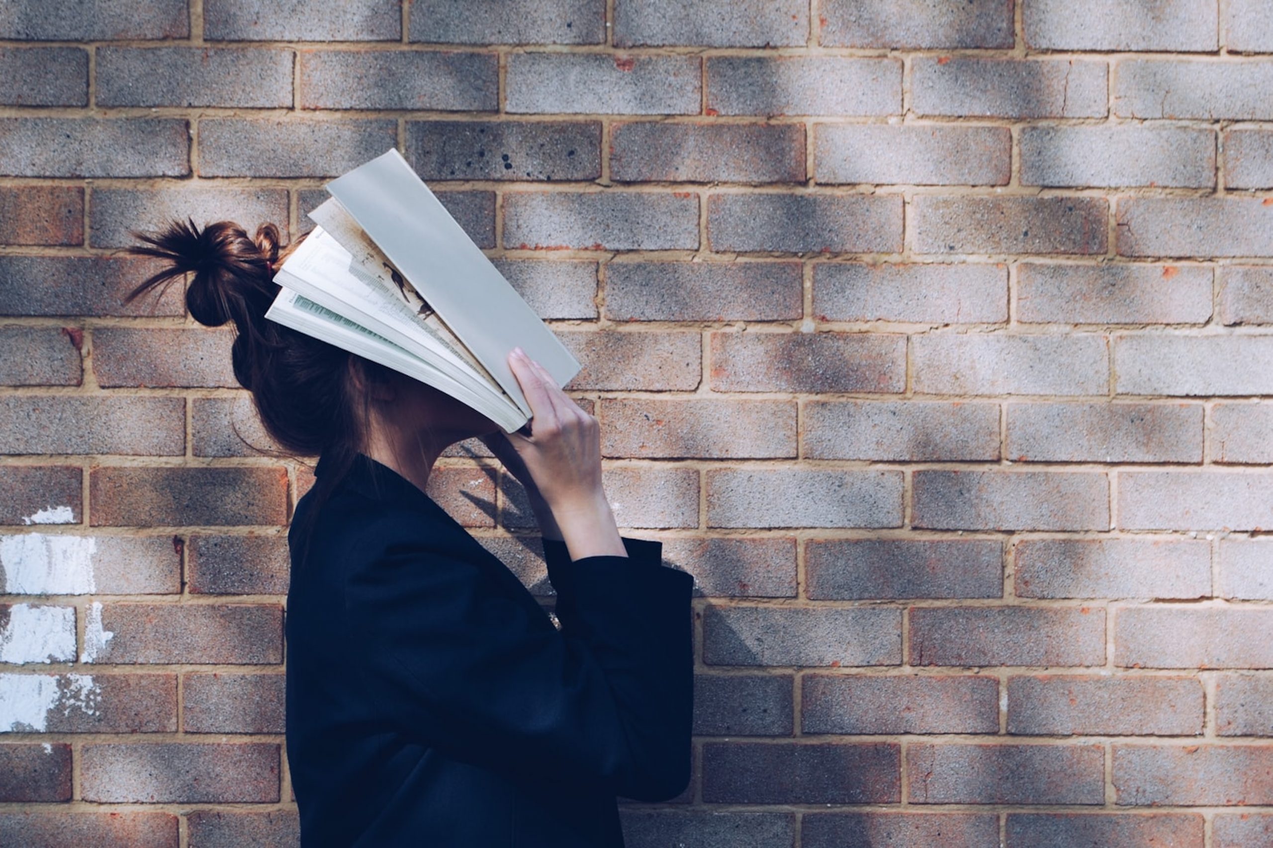 A girl in front of a brick all holding a book over her face.