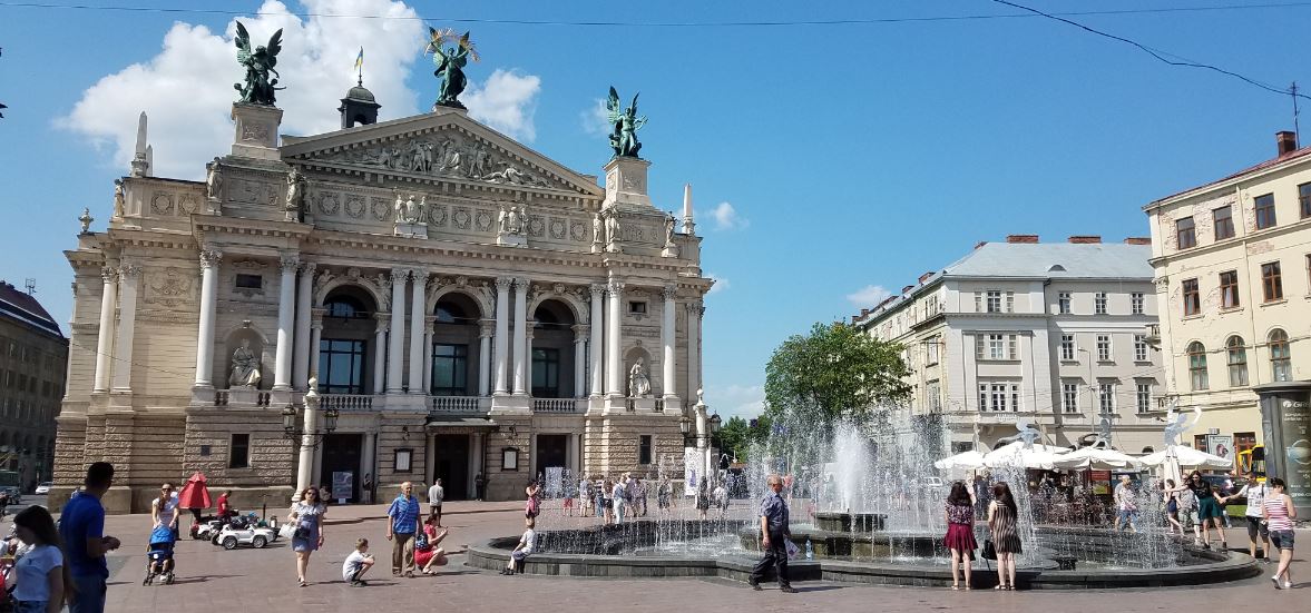 scene of square with fountain, statues and people of all ages. Large buildings surround the square and there is a cafe with umbrellas to one side.