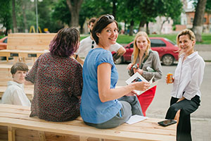 a group of people are siting, and standing, around a wooden bench. There is a woman looking back towards the camera while holding a booklet and smiling.