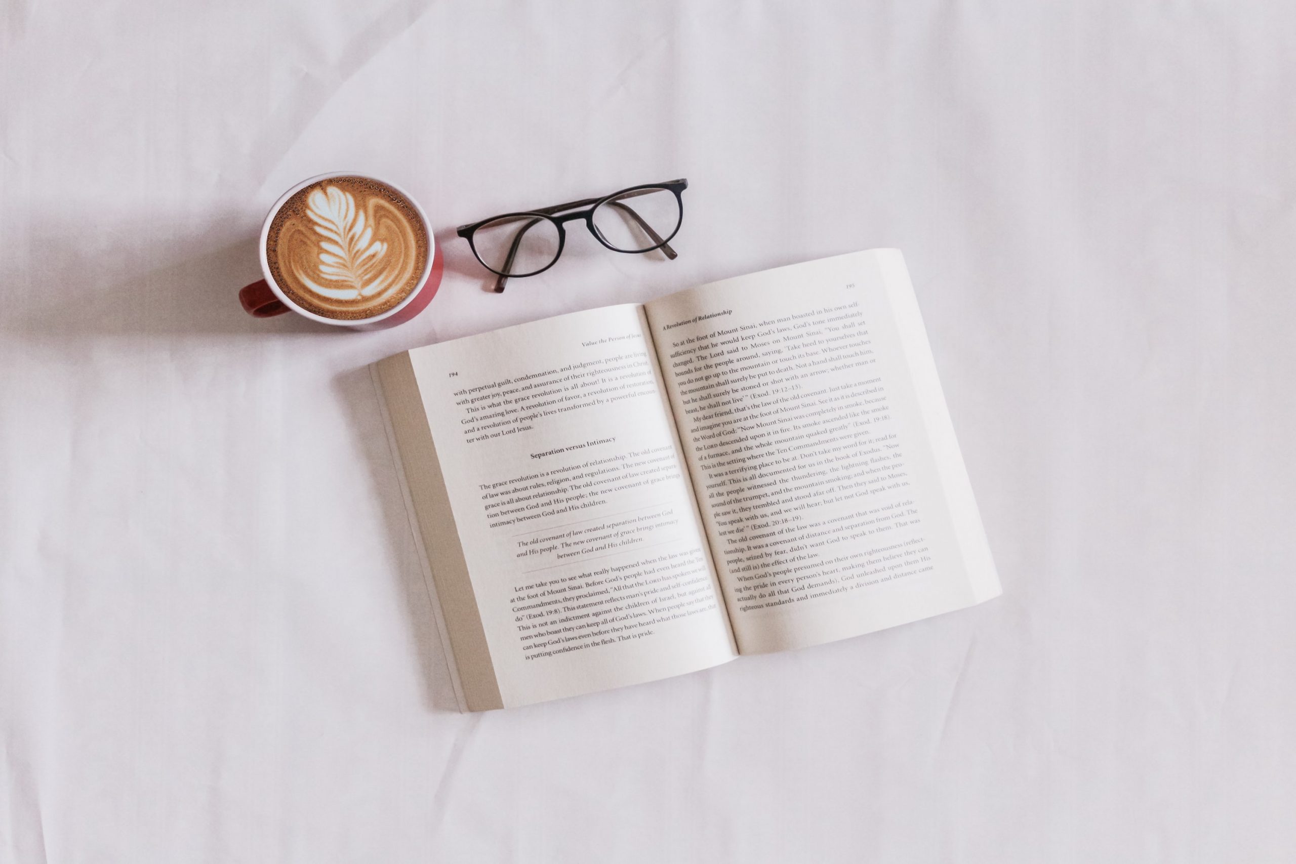 The imge is captured from a top view and everything is placed on top of a white cloth. An open book is parallel to a pair of glasses and a cup of coffee.