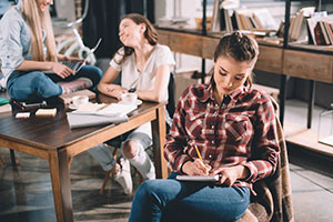 young woman writing in notebook with two girls talking behind her