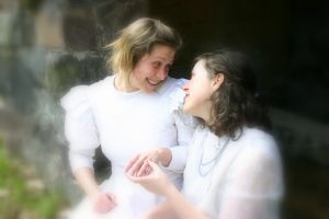 Lesbian couple in white dresses peer into each other's eyes and admire one's wedding ring.