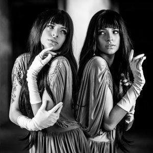 Black and white photo of two identical dark haired women, back-to-back, wearing dress gloves.