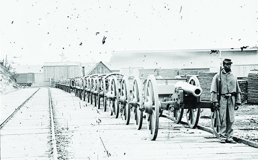 A photograph shows an African American soldier standing in front of a long line of cannons alongside a railroad track.