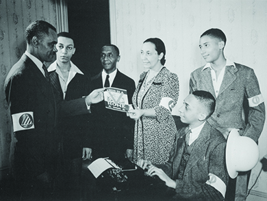 A photograph shows five Black men and a Black woman participating in the Double V campaign. A young man sits at a typewriter, and the woman hands a man a pamphlet, the cover of which reads “This is a [Double V insignia] Home.” All wear armbands.