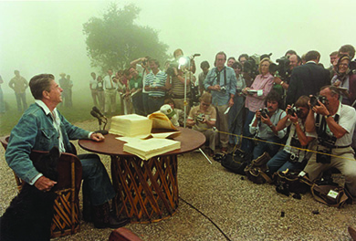 A photograph shows Ronald Reagan signing legislation while seated outdoors at a rustic table. He is dressed in blue jeans, a denim jacket, and cowboy boots, and he strokes the head of a large black dog seated beside him. In front of Reagan, the press takes photographs.